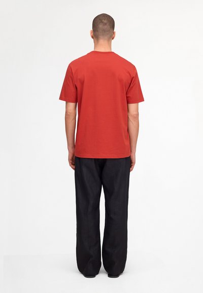 Man standing facing away, wearing a plain red short-sleeve t-shirt and loose black pants against a white background.