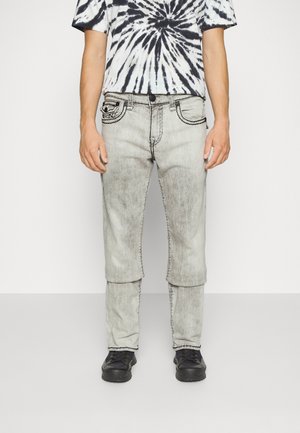 Homme portant un jean gris délavé avec des rabats horizontaux découpés au niveau des genoux et des baskets noires, associé à un t-shirt tie-dye noir et blanc.