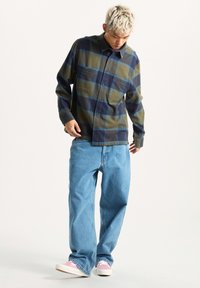 Camicia di flanella con motivo a quadri verde e blu navy; dettagli in teal, design con bottoni, due tasche sul petto. Abbinata a jeans azzurri a vestibilità ampia e scarpe rosa.