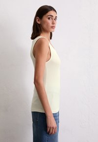 Lys gul ribbet singlet med brede stropper, med en tettsittende silhuett og sideskjæring, brukt med blå denimjeans.