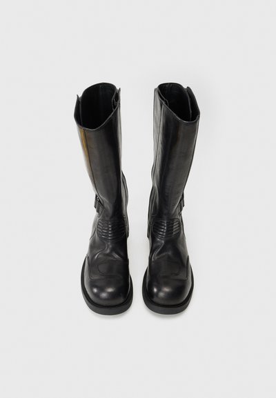 Cizme negre din piele până la genunchi, cu vârf rotund, accente laterale texturate și o talpă robustă din cauciuc. Au un finisaj neted și un design minimalist.