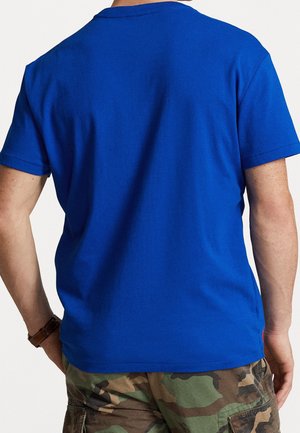 Person trägt ein schlichtes blaues T-Shirt mit kurzen Ärmeln und Tarn-Cargoshorts, von hinten gesehen.