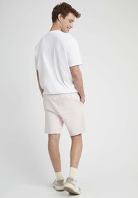 T-shirt en coton blanc associé à un short rose clair. Le short a une poche à l'arrière. Le modèle porte des chaussettes de couleur claire et des baskets.