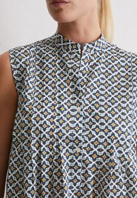 Kurzarmbluse aus Baumwolle mit geometrischem Muster in Marineblau, Weiß und Senfgelb. Verfügt über einen bandartigen Kragen und eine durchgehende Knopfleiste.