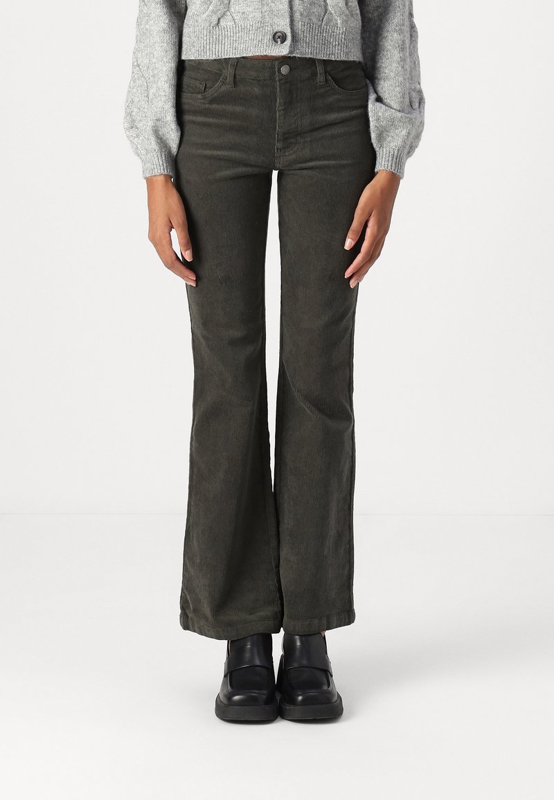 Femme portant un pantalon évasé en velours côtelé vert olive, un pull en tricot gris et des chaussures à plateforme noires, debout devant un fond blanc uni.