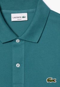 Turkusowa koszulka polo z teksturowanej tkaniny, wąski krój, trzy guziki przy kołnierzyku oraz zielone logo krokodyla po lewej stronie klatki piersiowej.