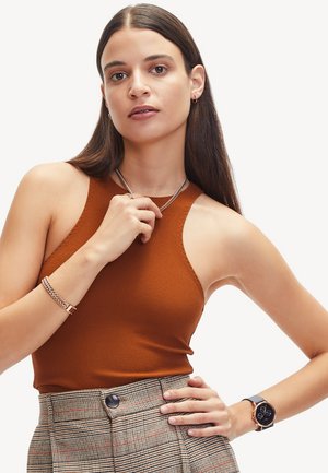 Haut sans manches marron, en tissu texturé, porté avec un pantalon à carreaux taille haute. Les accessoires comprennent un collier en chaîne argenté et une montre noire.