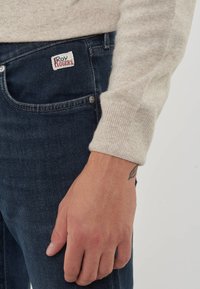 La mano destra di una persona che indossa un maglione beige, con la manica appoggiata vicino alla tasca dei jeans in denim blu con la patch del logo Roy Rogers.
