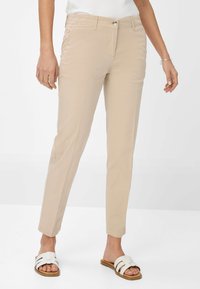 Pantalons beiges ajustés avec une texture lisse, devant plat et poches latérales, associés à des sandales blanches à enfiler avec plusieurs brides horizontales.