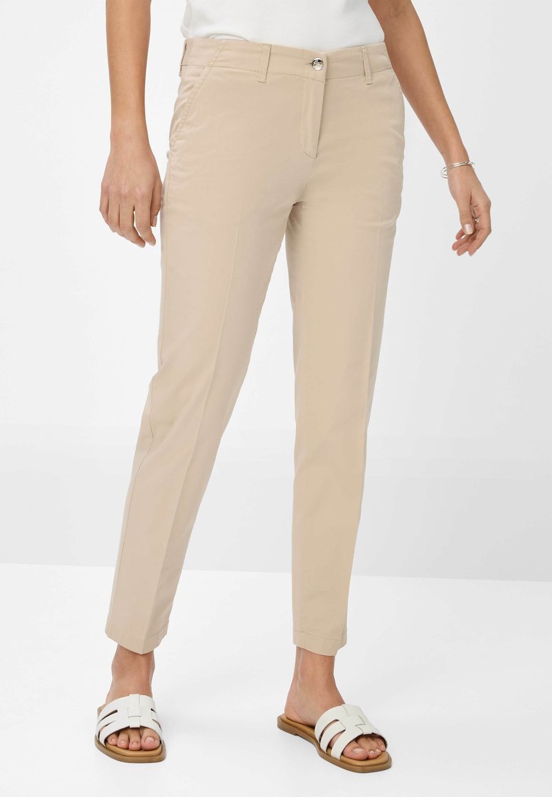 Pantalons beiges ajustés avec une texture lisse, devant plat et poches latérales, associés à des sandales blanches à enfiler avec plusieurs brides horizontales.