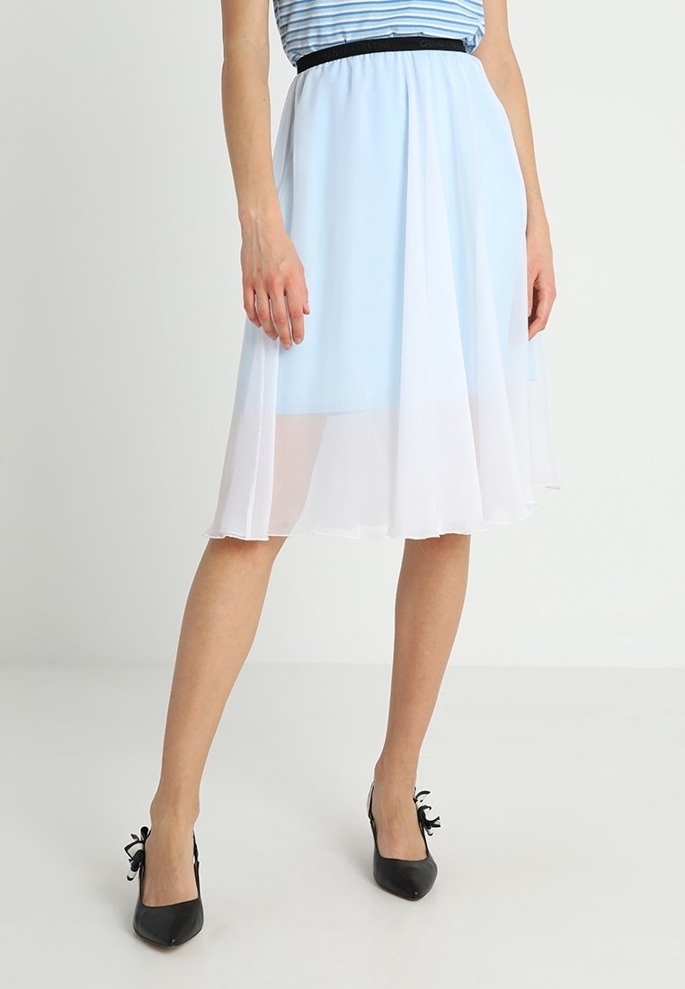 Femme portant une jupe midi bleu clair avec un voile blanc transparent et des chaussures à talons noires ornées de nœuds, debout devant un fond uni.