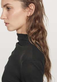 Black turtleneck top with a fitted texture, featuring a high neckline and ribbed detail. Model has wavy, shoulder-length brown hair.
