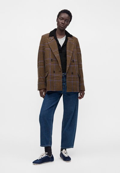 Femme portant un blazer à carreaux marron et bleu, une chemise noire, un sous-shirt blanc, un jean bleu ample, des chaussettes noires et des baskets bleu marine, debout sur fond blanc.