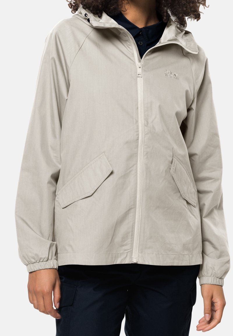 Personne portant une veste beige clair zippée avec capuche et poches à rabat par-dessus une chemise sombre et un pantalon foncé.