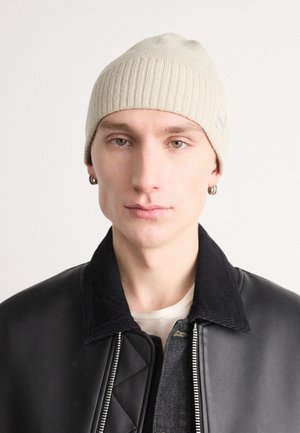 Jeune personne portant un bonnet beige en tricot, des boucles d'oreilles en argent en forme de créoles, une chemise blanche et une veste noire avec un col en velours côtelé, tournée vers l'avant.