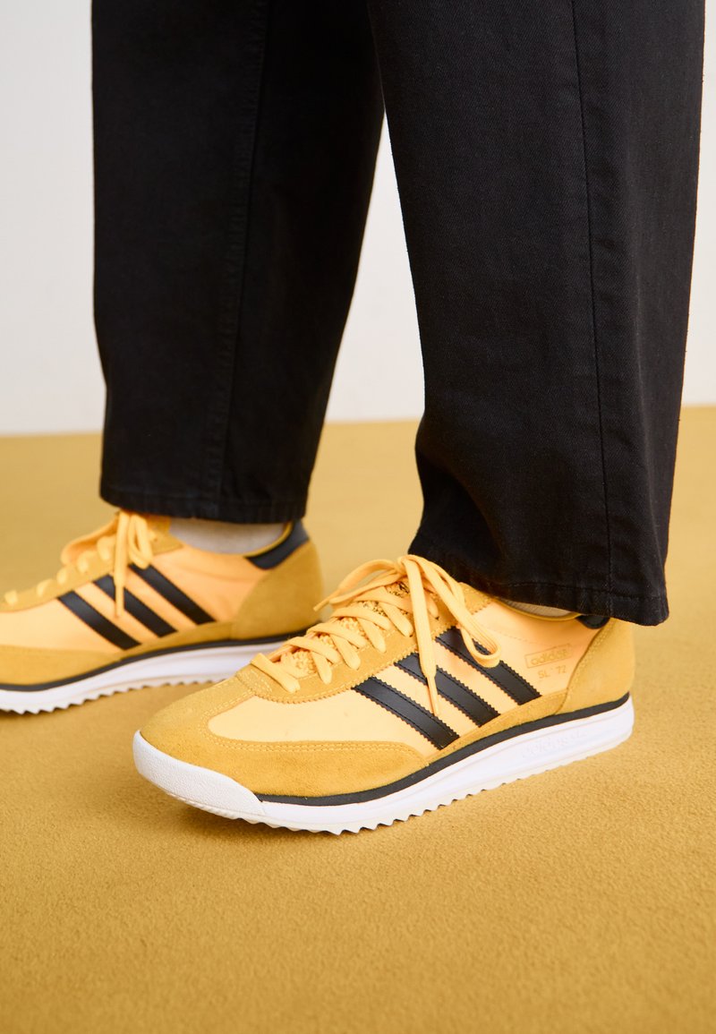 Gelbe Sportschuhe mit schwarzen Streifen, aus Wildleder und Mesh-Material, weißer Sohle und einem markanten Retro-Design, kombiniert mit schwarzen Hosen.