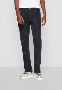 Black denim jeans with smiley face patterns, straight leg design, five-pocket style, and a green button accent. Worn with colorful patterned sneakers.