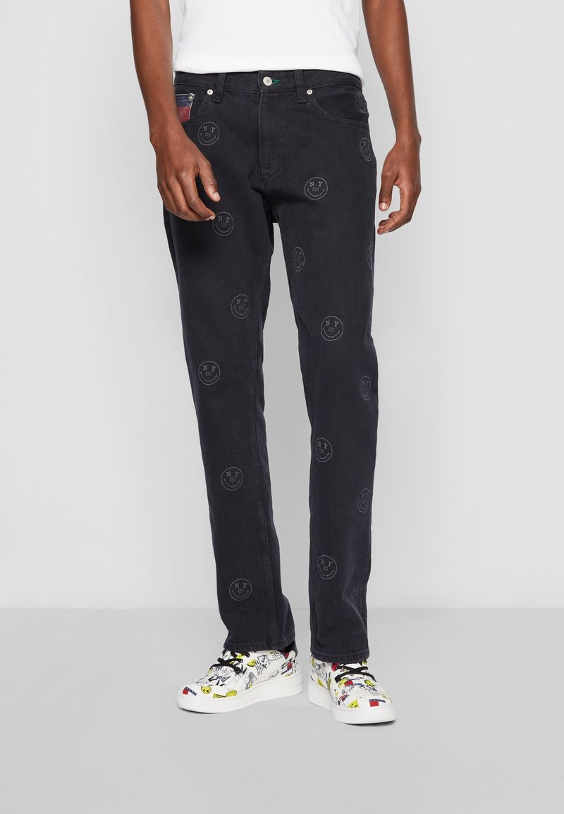 Black denim jeans with smiley face patterns, straight leg design, five-pocket style, and a green button accent. Worn with colorful patterned sneakers.