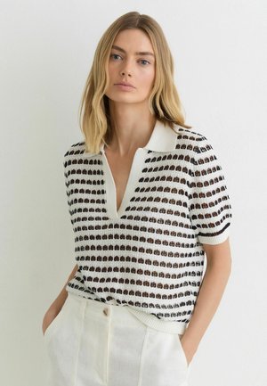 Blondynka w białej, krótkim rękawem, dzianinowej bluzce z czarno-brązowymi wzorami oraz białych spodniach z wysokim stanem, pozuje z rękami w kieszeniach.