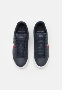 Sneaker in pelle blu scuro con suole in gomma bianche, caratterizzate da un design con lacci e strisce rosse e bianche sui lati. Logo Tommy Hilfiger sulla linguetta.