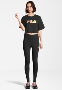 Camiseta negra cortada con un colorido logo de "FILA", combinada con leggings negros ajustados. Modelo lleva zapatillas negras con acentos rosas.