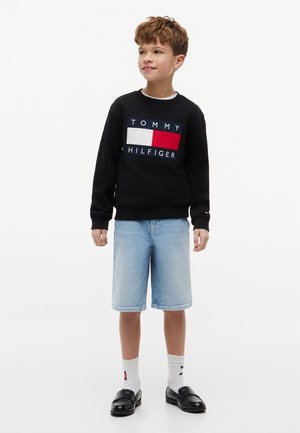Chłopiec w czarnej bluzie Tommy Hilfiger, jasnoniebieskich szortach z denimu, białych skarpetach i czarnych mokasynach, stoi i patrzy lekko w prawo.