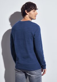 Man in een blauwe langemouwen shirt en grijze spijkerbroek staat met zijn gezicht naar een witte muur, kijkt over zijn rechter schouder.