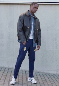 Zwarte leren jas over een spijkerbroekhemd, wit T-shirt, marineblauwe joggingbroek met een geel embleem, en witte sneakers met zwarte strepen.