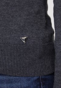 Grå stickad tröja med ribbad nederkant och liten metallisk triangel-logotyp på sidan, med ett mjukt, strukturerat tyg.