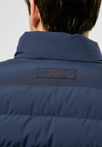 Hombre con chaqueta acolchada de color azul oscuro y cuello alto, mostrando la parte posterior con una pequeña etiqueta cosida que dice "MCS" en hilo rojo.