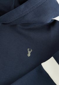 Sweatshirt en coton bleu marine avec un logo de cerf argenté, des poignets côtelés et une bande blanche contrastante le long des manches.