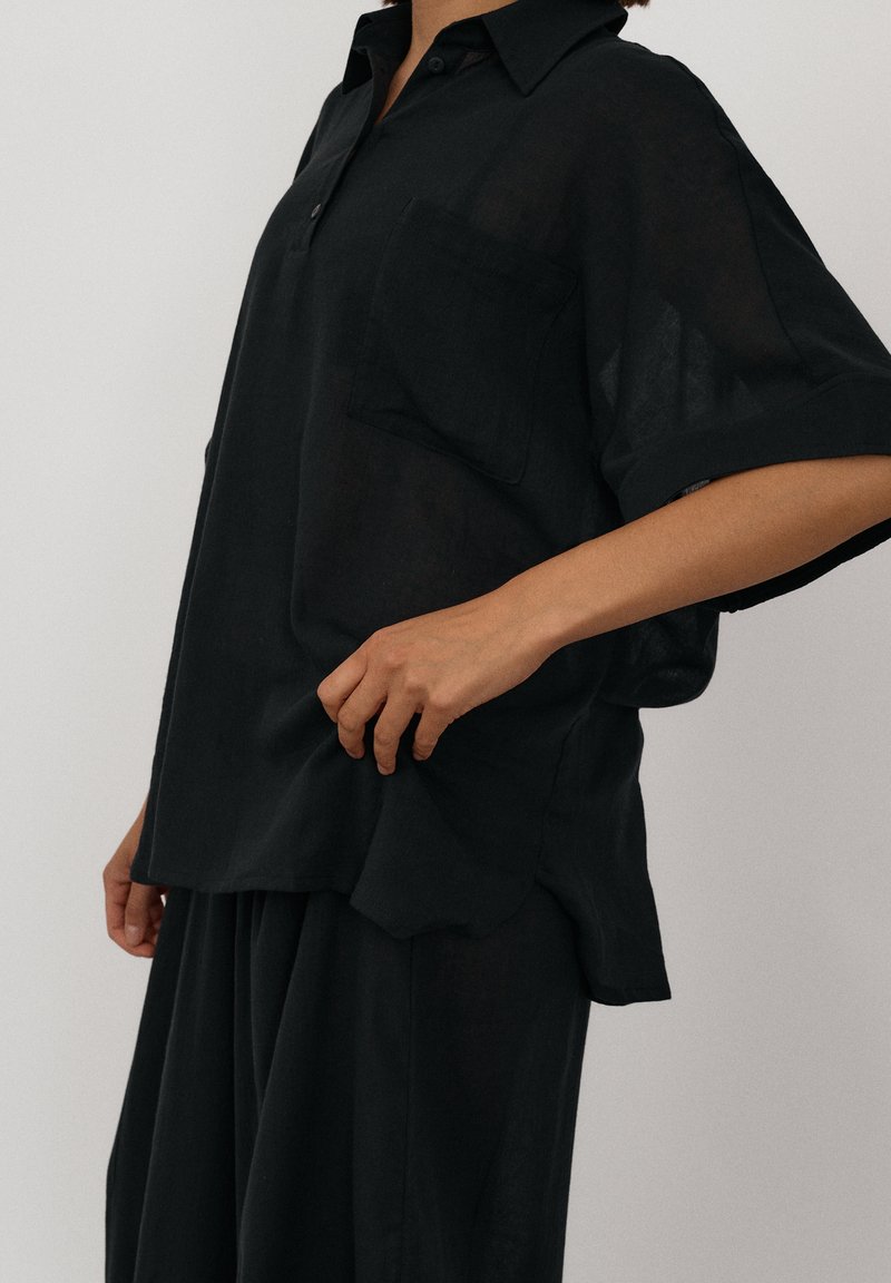 Osoba ubrana w luźną czarną koszulę z krótkim rękawem i kieszenią oraz pasujące szerokie spodnie, stojąca na tle jednolitego jasnego tła.