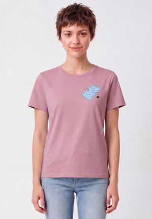 Jeune personne aux cheveux courts portant un t-shirt rose avec un petit motif piscine sur la poitrine et un jean bleu clair.