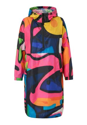 Kvinde iført en lang jakke med farverigt abstrakt mønster og hætte, blå opsmøgede jeans og gule sneakers, stående mod en hvid baggrund.