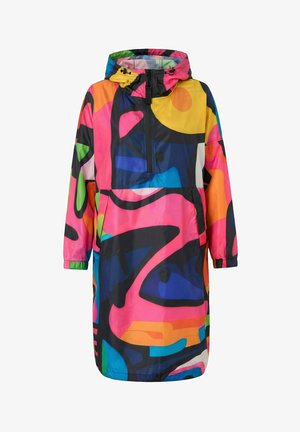 Nainen, jolla on päällään värikäs abstraktikuosinen pitkä hupparitakki, siniset käännetyt farkut ja keltaiset tennarit, seisoo valkoista taustaa vasten.