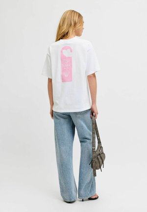 Femme aux longs cheveux blonds portant un t-shirt blanc avec un motif rose « Please Knock » sur un accroche-porte dans le dos, jean bleu clair, tenant un sac gris.