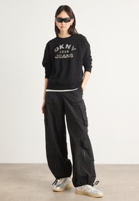 Svart hettegenser med "DKNY 1989 JEANS" logo, kombinert med løse svarte cargo-bukser og sølvfargede sneakers; modell med svarte solbriller.