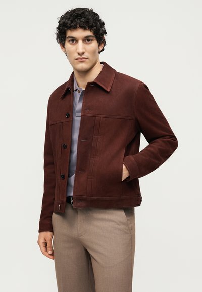 Νέος άνδρας με σγουρά μαύρα μαλλιά, φορά καφέ κουμπωτό σακάκι πάνω από γκρι πουκάμισο και μπεζ παντελόνι, στέκεται με το ένα χέρι στην τσέπη.