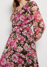 Kvinna klädd i en genomskinlig långärmad blommig klänning i rosa, vitt och grönt med V-ringning och knytbart midjebälte.