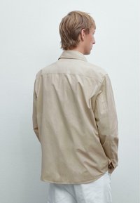 Ung person med kort blond hår iført en beige langærmet skjorte og hvide bukser, set fra ryggen mod en ensfarvet væg.