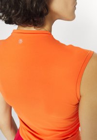 Femme aux cheveux courts et bouclés portant un débardeur de sport orange vif, vue de dos sur un fond blanc uni.