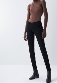 Pantalones ajustados negros con cintura alta y cierre de tres botones, combinados con una blusa marrón de un solo hombro de canalé y botas de tacón negras hasta el tobillo.