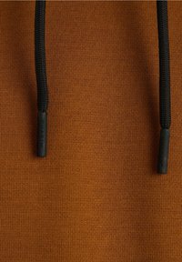 Gros plan d’un tissu marron texturé avec deux cordons noirs, chacun terminé par une pointe en plastique noir embossée du mot "CHASIN".