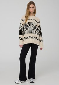 Maglione oversize lavorato a maglia in crema con motivi geometrici neri, abbinato a pantaloni svasati neri e sneakers argento. Design testurizzato e casual.