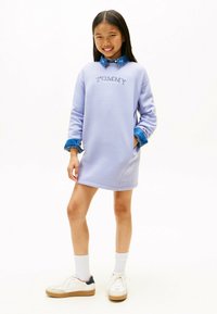 Lilla sweatshirt-kjole med et broderet logo, opkrøllede denimærmer og en behagelig pasform, parret med hvide sneakers og knæhøje sokker.