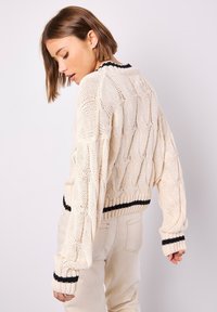 Cremfarbener Kabelstrickpullover mit einem cropped Design, ausgestattet mit schwarz gestreiften Bündchen und Saum. Der Stoff hat eine weiche, texturierte Oberfläche.