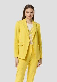 Blazer giallo su misura con rever a punta, chiusura con un bottone e due tasche frontali, abbinato a pantaloni coordinati e una T-shirt bianca con grafica.