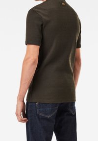 Donkergroen geribbeld polo shirt met korte mouwen, heeft een klein label op de achterkant nabij de halslijn, gecombineerd met donkerblauwe jeans.