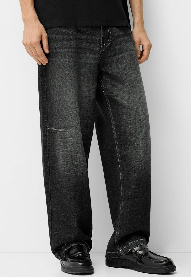 Homme portant un pantalon en jean large de couleur foncée avec des coutures visibles et des mocassins en cuir noirs, adossé à un mur blanc.