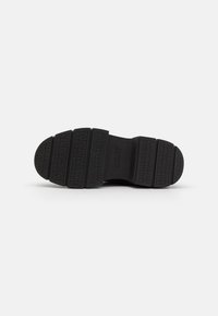Semelle de chaussure en caoutchouc noir avec un motif de semelle prononcé, featuring le nom de marque embossé "DKNY" et des accents rectangulaires texturés.